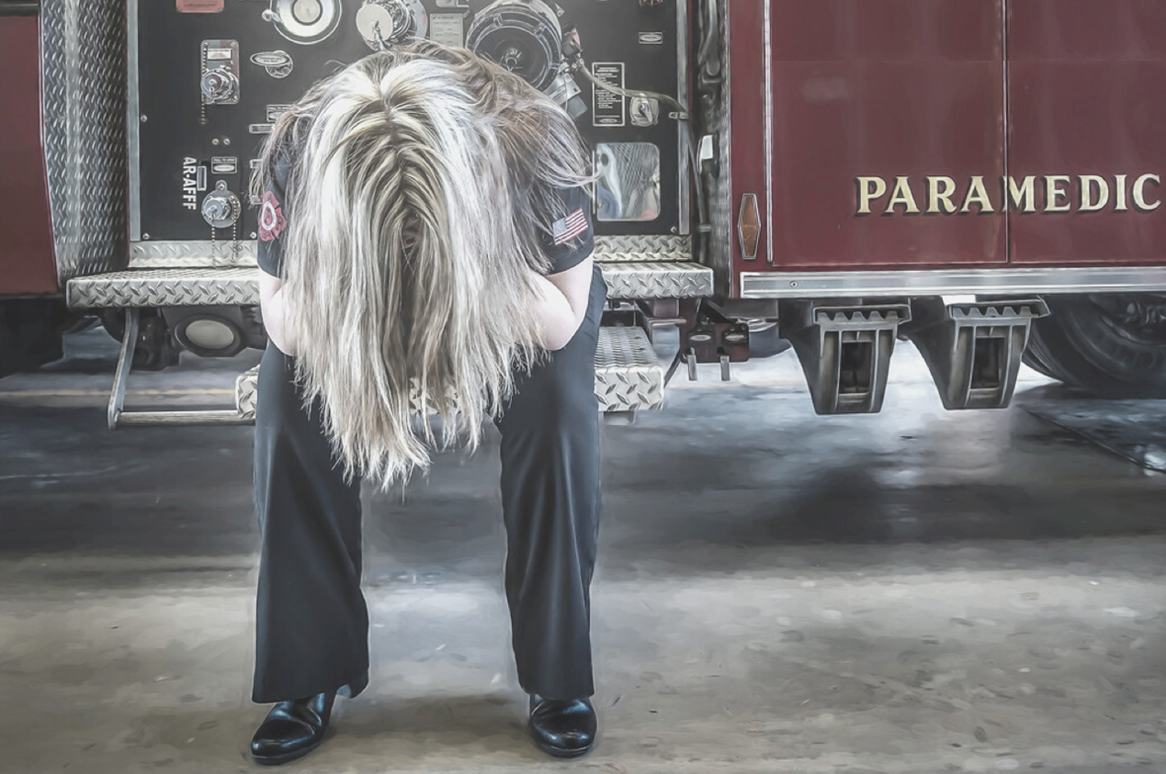 A picture of a flre fighter sitting on a fire truck, holding her head in her hands.