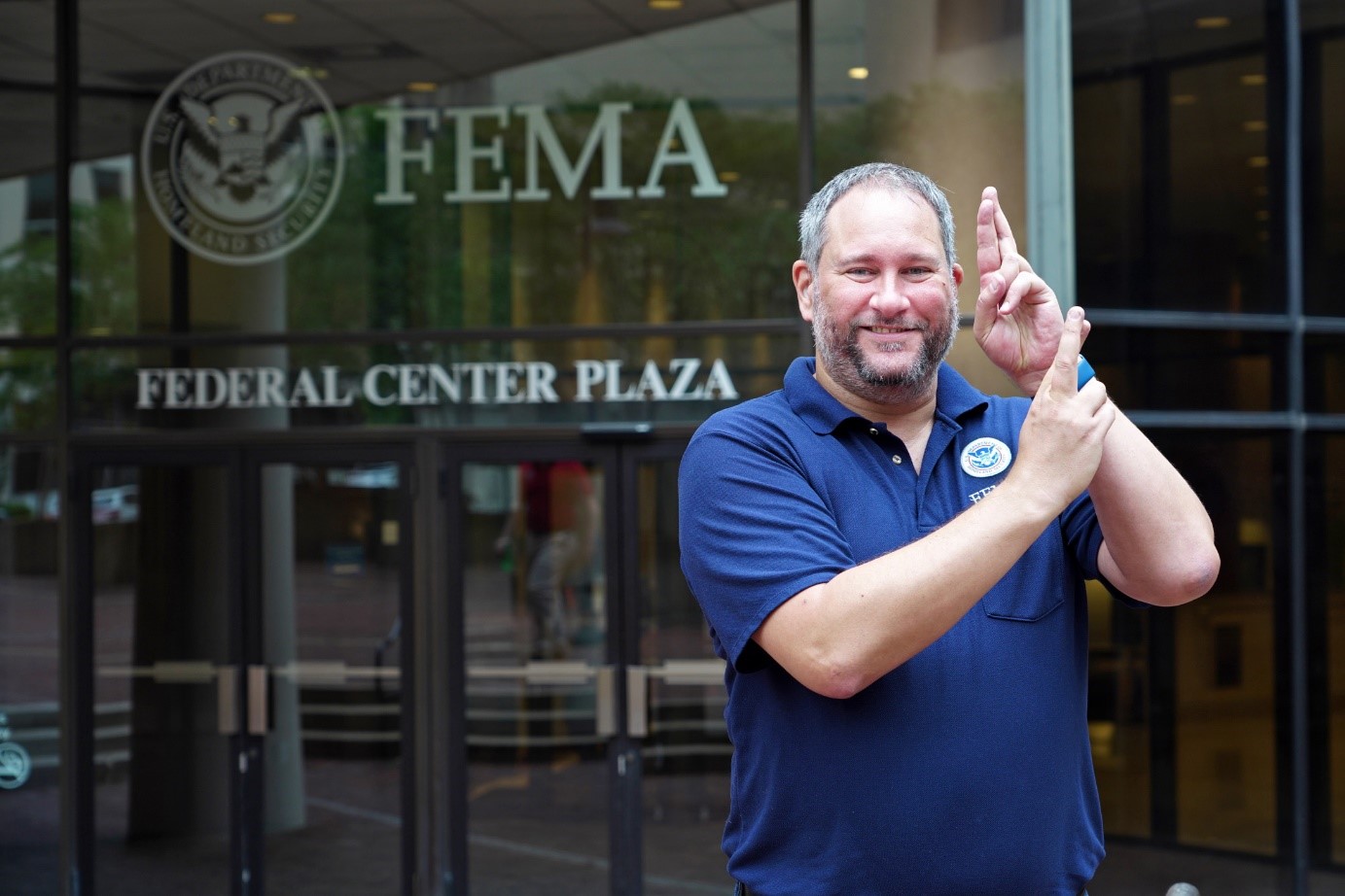 Aaron Kubey stands in front of a glass door at FEMA head quarters, wearing a blue polo shirt. 