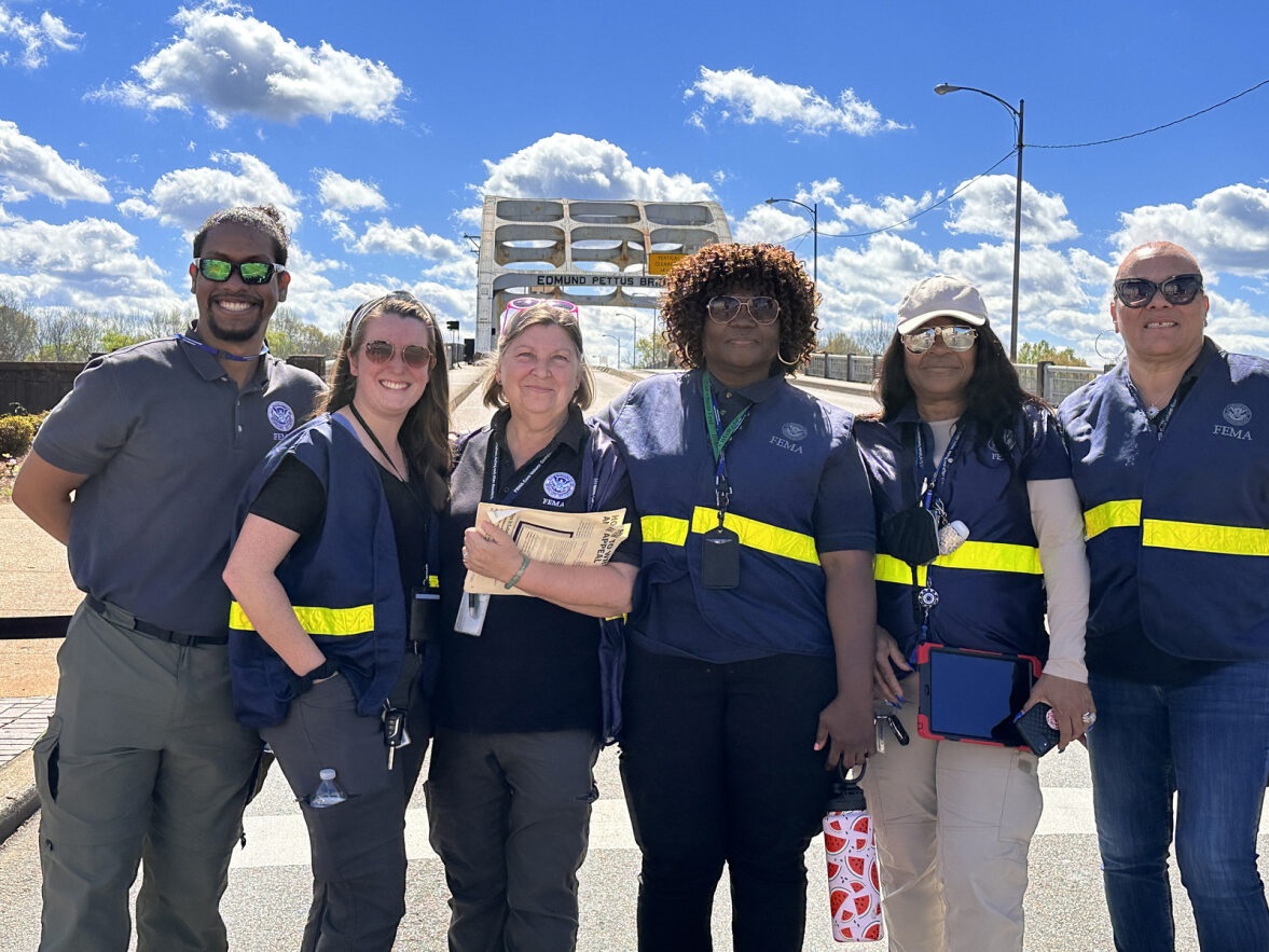 Six FEMA employees in bright safety vests smile at outdoor work site.