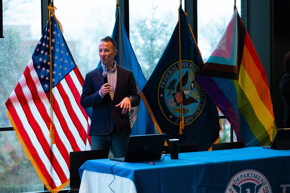 Man with microphone speaks in front of backdrop of flags